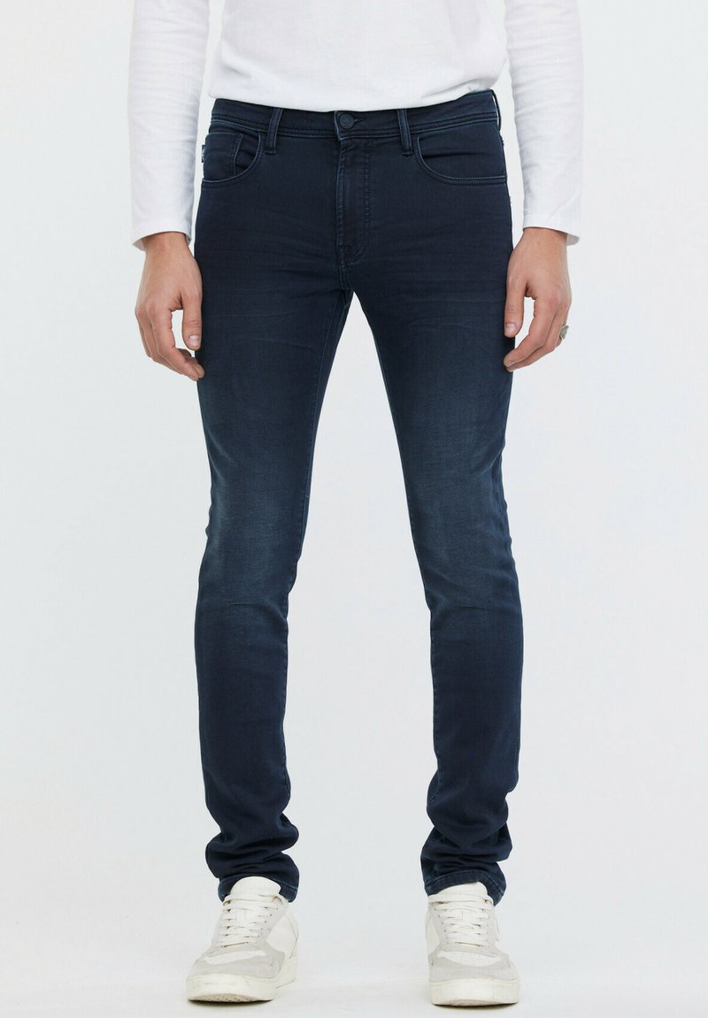Jean skinny bleu foncé en denim. Dispose de poches avant, passants de ceinture et d'une fermeture à bouton. Porté avec des baskets blanches.