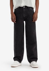 Persona vestida con pantalones vaqueros anchos negros y zapatillas blancas, de pie contra un fondo blanco liso.
