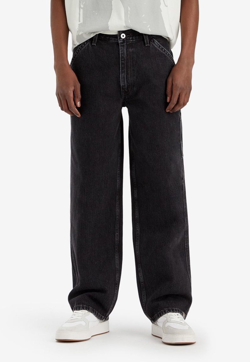 Persona vestida con pantalones vaqueros anchos negros y zapatillas blancas, de pie contra un fondo blanco liso.