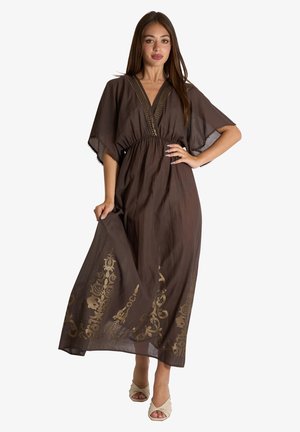 Donna che indossa un abito maxi marrone con ricami floreali dorati sull'orlo e maniche larghe, in piedi con una mano sul fianco e l'altra che tiene il vestito.