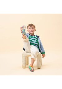 Sandalias coloridas con una base verde, correas estampadas y una suela multicolor. Chaqueta de punto azul claro, camisa a rayas y pantalones blancos.