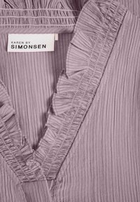 Blouse à col en V en tissu texturé lavande avec des accents volantés le long de l'encolure, portant une étiquette indiquant "KAREN BY SIMONSEN".