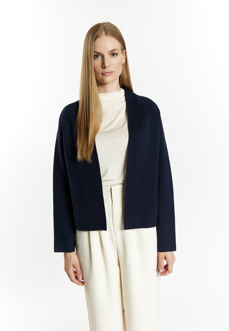 Marineblauwe cropped cardigan gemaakt van een textuurstof, met een brede open voorkant, lange mouwen en een relaxte pasvorm, gecombineerd met een licht beige top.