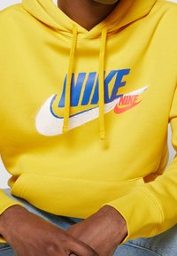 Person som bär en gul hoodie med blå och röda Nike-logotyper, med händerna ihopfällda och vilande på ljusblå jeans.