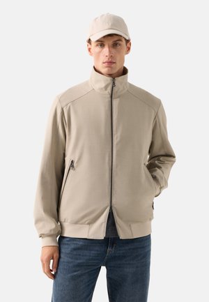 Junger Mann trägt eine beige Reißverschlussjacke mit Stehkragen, passende Mütze und blaue Jeans, steht vor einem schlichten hellen Hintergrund.