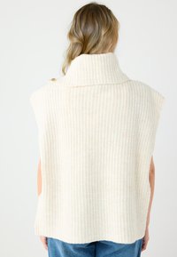 Gilet in maglia color crema con collo alto a coste, design senza maniche e spacco sui lati. Presenta un dettaglio con bottone color oro sul collo.