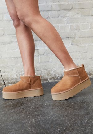 Personne portant des bottines en daim beige avec des semelles épaisses beige clair, debout sur un sol en béton contre un mur en briques claires.