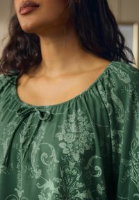 Zaļš blūze ar ziedu rakstu, apaļa izgriezuma, savilktiem plecu šuvēm un dekoratīvu saišu priekšpusē.