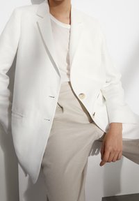 Biały lniany żakiet zapinany na jeden guzik, z klapami w kształcie notcha i dwoma przednimi kieszeniami; zestawiony z jasnobeżowymi spodniami. Prosty i dopasowany krój.