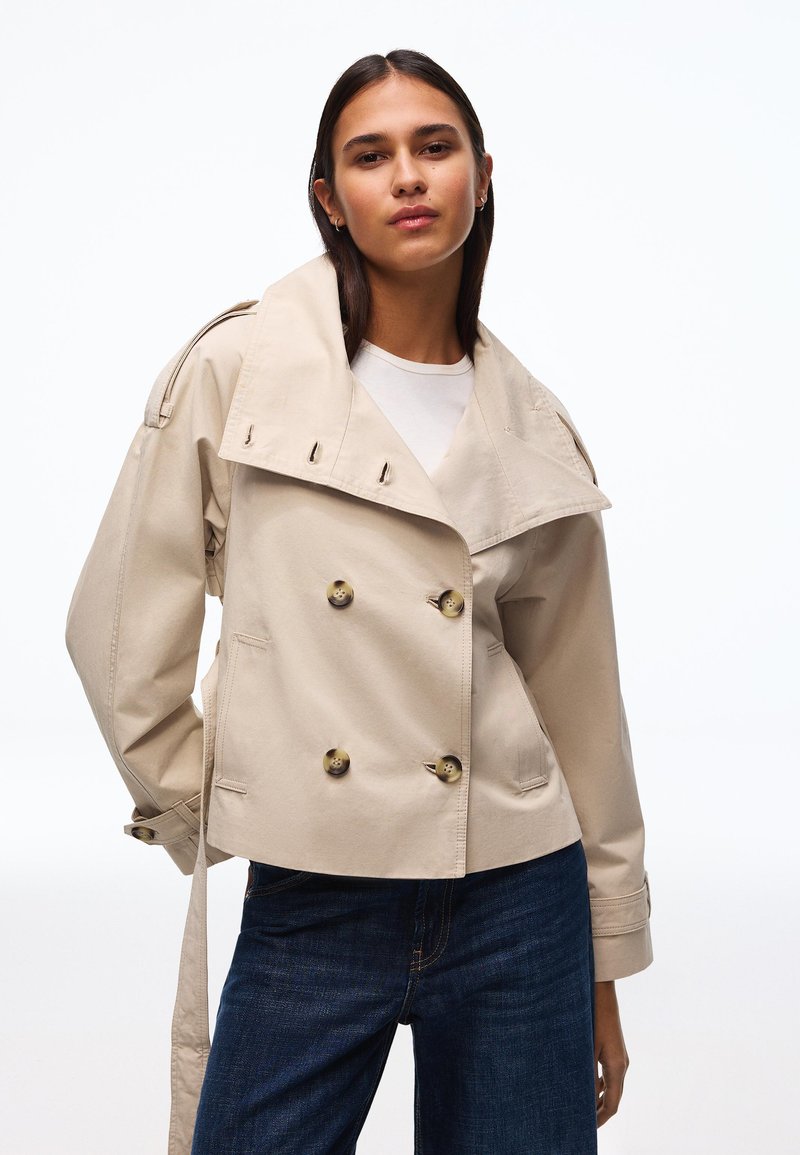 Junge Frau trägt beige zweireihigen Trenchcoat über weißem Oberteil und dunkelblauen Jeans, steht vor weißem Hintergrund.