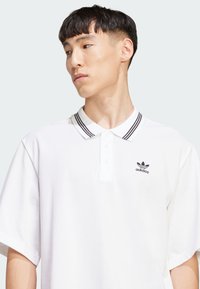 Witte polo met een geribbelde kraag met zwarte strepen en een geborduurd zwart Adidas-logo op de borst. Korte mouwen.