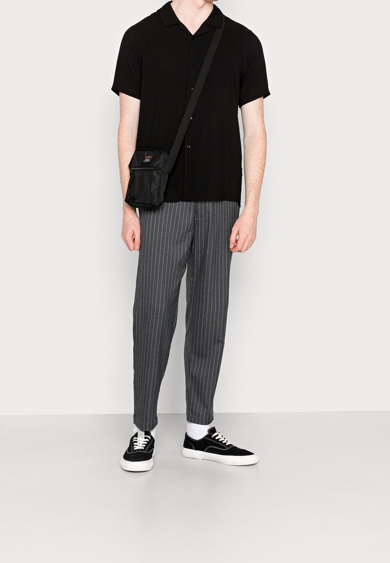 Homme portant une chemise noire à manches courtes boutonnée, un pantalon gris à fines rayures, des baskets noires et blanches, des chaussettes blanches et un sac en bandoulière noir.