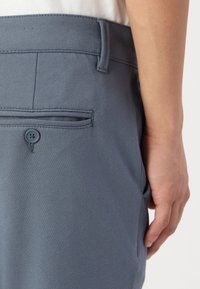 Los pantalones grises presentan una textura suave, bolsillos estándar y un cierre de botón en la parte posterior. La cintura está cuidadosamente cosida con un detalle mínimo.