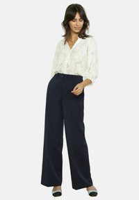 Blusa blanca con estampado floral, cuello en V y mangas largas, combinada con pantalones de talle alto, de pierna ancha en azul marino, y zapatos negros y azules.