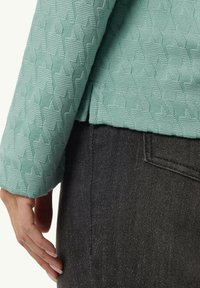 Helle türkisfarbene, strukturierte Jacke mit geometrischem Muster, kombiniert mit dunklen Denim-Hosen. Nahaufnahme zeigt Details des Ärmels.