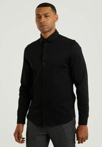 Camisa negra de manga larga con botones en la parte delantera, tela sólida y acentos en los hombros. Presenta un pequeño logo en el pecho.