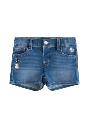 Ung pige iført en hvid t-shirt med sommerfugleprint, denimshorts, en lavendelfarvet kasket og sandaler med leopardprint, stående på en ensfarvet baggrund.