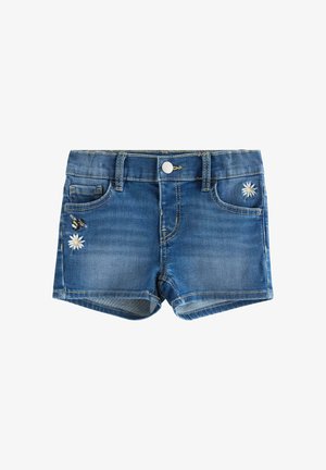 Ung pige iført en hvid t-shirt med sommerfugleprint, denimshorts, en lavendelfarvet kasket og sandaler med leopardprint, stående på en ensfarvet baggrund.