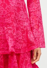 Φωτεινό ροζ φλοράλ φόρεμα με δαντέλα, μακριά μανίκια και στρωτή φραμπαλά στο τελείωμα, που δείχνει μέρος του χεριού ενός ατόμου στο πλάι.