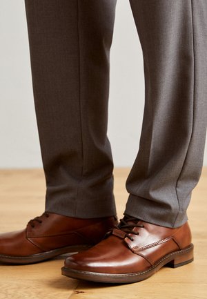Brown leather shoes with a smooth finish, lace-up design, and low heels, paired with gray, tailored trousers, featuring a slight sheen.