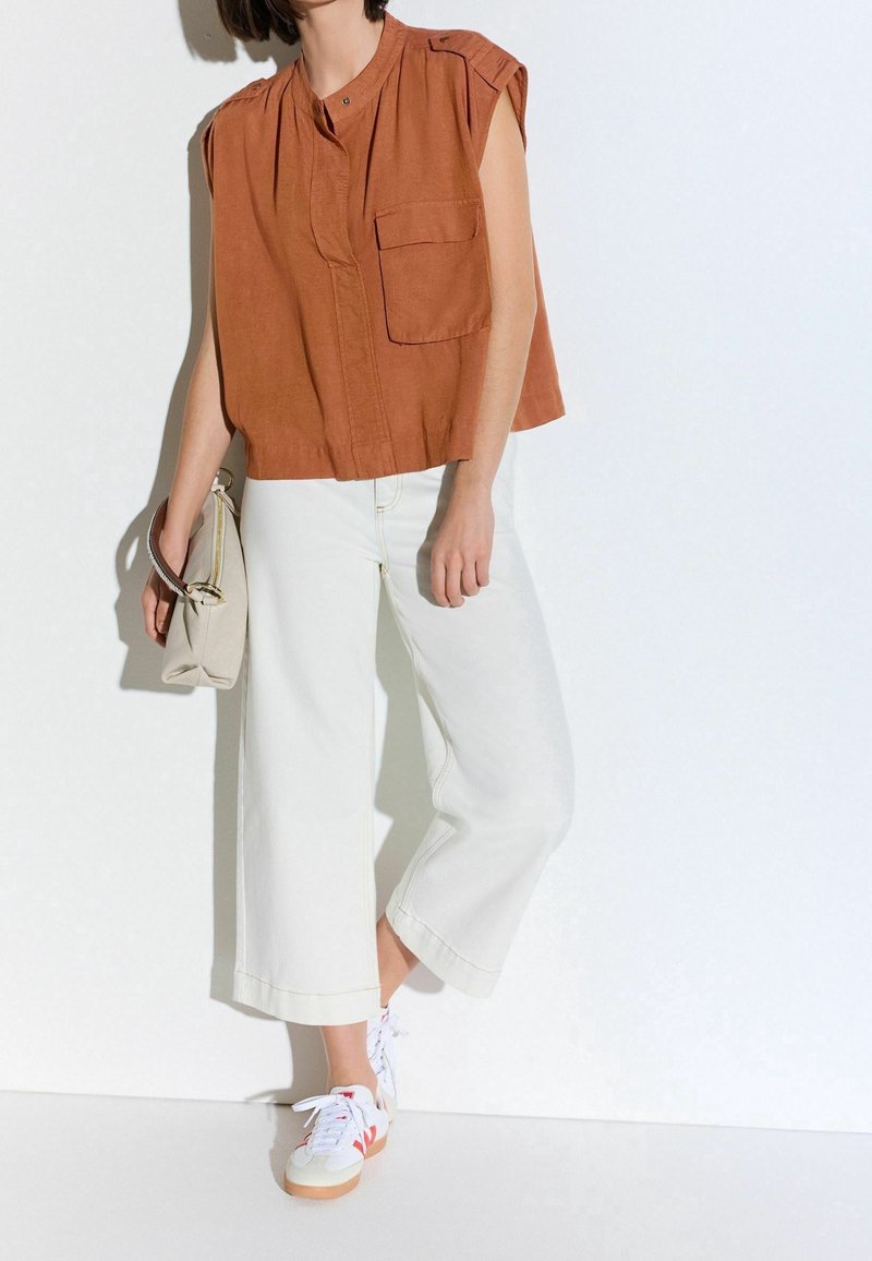 Personne portant un haut sans manches couleur rouille, pantalon large blanc, baskets blanches, et tenant un sac à main beige contre un mur blanc.