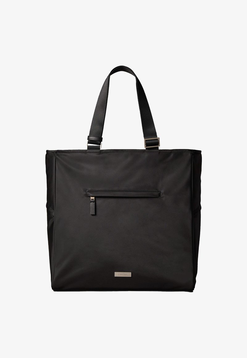 Μαύρη ορθογώνια τσάντα ώμου με διπλούς ιμάντες, εμπρόσθια τσέπη με φερμουάρ και μικρή μεταλλική πλακέτα με λογότυπο στο κάτω κέντρο.