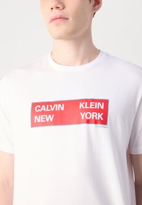 Balta kokvilnas T-kreklus ar izteiksmīgu sarkanu taisnstūra grafiku, kurā ir teksts "CALVIN NEW KLEIN YORK", kā arī neliels "Calvin Klein" logo.