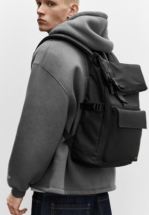 Mladý človek v sivej mikine s kapucňou, ktorý nosí čierny batoh s predným vreckom a zapínaním na pracku, pozerajúci sa cez plece na jednoliatom pozadí.