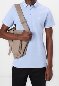 Polo shirt azzurro chiaro con rifiniture bianche, colletto, abbinato a una borsa a tracolla beige con tasche con zip e spallacci piatti. Pantaloni neri.