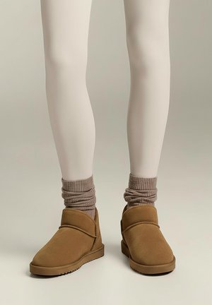 Beine mit hellbeigen Strumpfhosen, braunen lässigen Socken und braunen Slip-On-Stiefeletten mit flachen Sohlen vor einfachem Hintergrund.