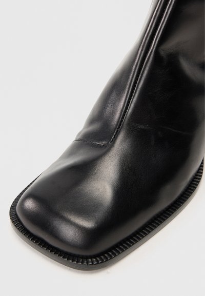 Κοντινό πλάνο ενός μαύρου δερμάτινου μποτακιού με τετράγωνο τακούνι και λεπτομερείς ραφές στην άκρη της σόλας σε λευκό φόντο.