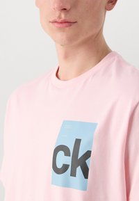 Rozā kokvilnas t-krekls ar lielu zilu kvadrātu ar melnu uzrakstu. Uzraksti ietver "calvin klein jauno jorku" un "kopš 1968. gada."