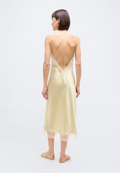 Γυναίκα με κοντά σκούρα μαλλιά που φοράει κρεμ σατέν φόρεμα με δαντέλα στη λεπτομέρεια, εμφανίζεται ξυπόλητη από την πίσω πλευρά με φόντο ένα απλό λευκό φόντο.