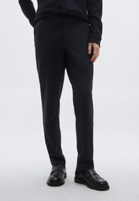 Na mieru šité čierne nohavice z hladkej látky, so strihom s rovnými nohavicami, bočnými vreckami a v kombinácii s čiernymi mokasínmi.