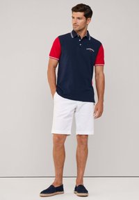 Camisa polo color bloque en azul marino y rojo con cuello blanco y acentos a rayas, combinada con pantalones cortos blancos y espadrilles en azul marino.