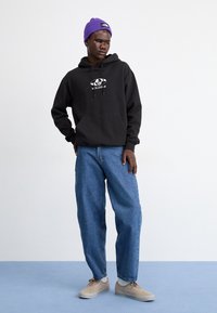 Homme portant un hoodie noir Volcom, un jean bleu ample, des baskets beiges, un bonnet en tricot violet, se tenant sur un sol bleu devant un mur blanc uni.