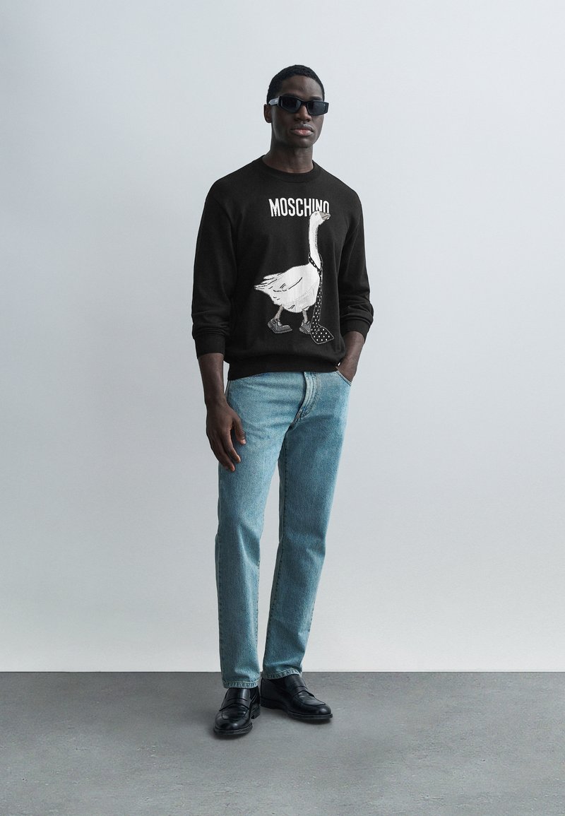 Schwarzer Pullover mit weißem Entenmotiv und Punkten auf den Schuhen, kombiniert mit hellblauen Jeans und schwarzen Schuhen, vor einem einfarbigen Hintergrund.