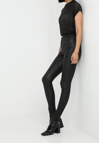 Pantalones de cuero negro de cintura alta combinados con una blusa negra transparente y plisada. La modelo lleva botas de tacón negras brillantes.