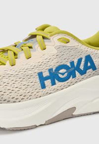 Könnyű futócipő, bézs és sárga akcentusokkal ellátott hálós felsőrésszel. Kék "HOKA" logóval és texturált gumi talppal rendelkezik.
