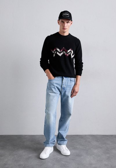 Svart stickad tröja med ett zigzag-mönster i vinröd och krämfärgad, kombinerad med ljusblå jeans och vita sneakers. Svart keps med textdetalj.
