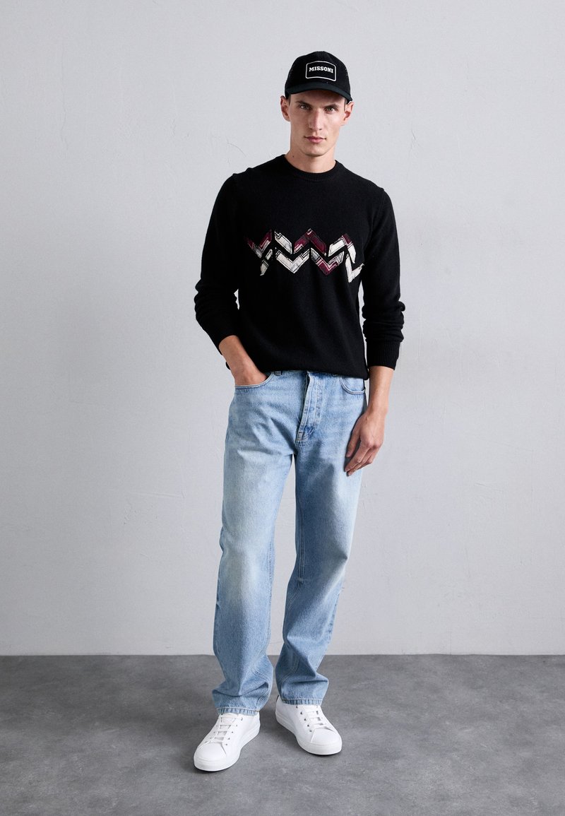 Schwarzer Strickpullover mit einem Zickzackmuster in Burgundy und Creme, kombiniert mit hellblauen Jeans und weißen Sneakern. Schwarze Mütze mit Textakzent.