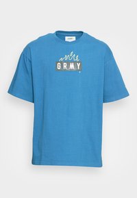 T-shirt en coton bleu avec une coupe décontractée, ornée d'un graphisme rectangulaire imprimé avec le texte "GRMY" et un design de flamme jaune au-dessus.
