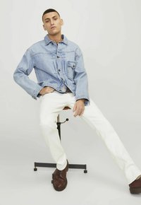 Giacca di jeans di colore azzurro chiaro con chiusura a bottoni e tasca sul petto, abbinata a pantaloni bianchi e scarpe in pelle marrone, seduto su uno sgabello.