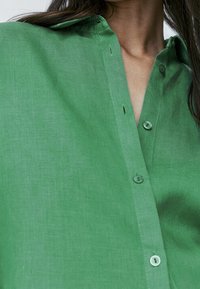 Woman wearing a green textured long-sleeve shirt with buttons, showing part of her neck and collarbone.