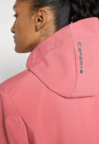 Femme aux cheveux foncés attachés porte une veste à capuche rose avec un texte de marque visible à l'arrière de la capuche.