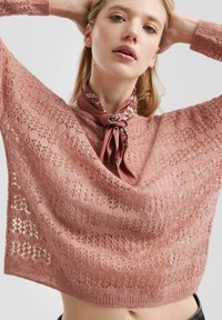 Junge Frau, die einen rosa Spitzenstrickpullover und einen gemusterten braunen Halstuch trägt, posiert mit den Händen hinter dem Kopf vor einem schlichten Hintergrund.