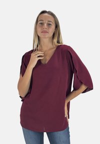Blusa bordeaux con scollo a V, maniche corte svasate, realizzata in tessuto leggero, caratterizzata da una texture liscia e una vestibilità rilassata.