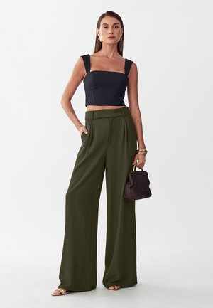 Mujer de pie con un top corto negro, pantalones anchos de color verde oliva, pendientes dorados, pulseras, sosteniendo un pequeño bolso tejido.