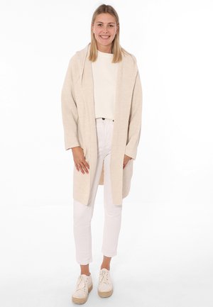 Hellbeige offene Cardigan mit langen Ärmeln, strukturierter Stoff, kombiniert mit einem weißen Oberteil und einer weißen Hose. Getragen mit weißen Espadrilles.