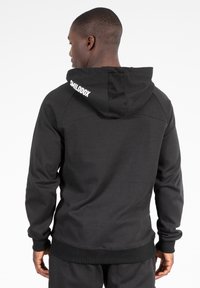 Černá mikina s kapucí vyrobená z měkké tkaniny, vybavená stahovací šňůrkou a žebrovanými rukávy. Logo "SMILODOX" v bílé barvě na horní části zad.